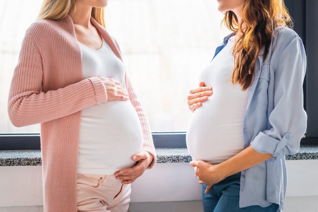 cropped view of smiling pregnant women standing by window